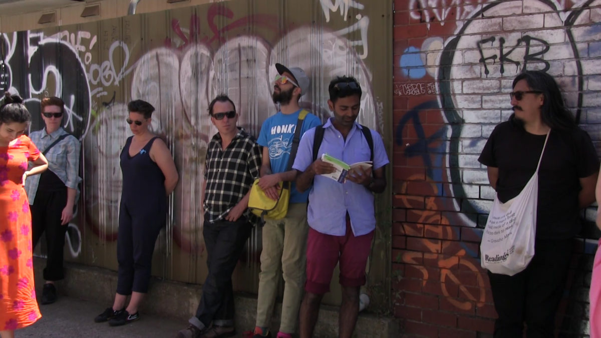 A group of people in front of a graffitied wall. One person reads from a book.