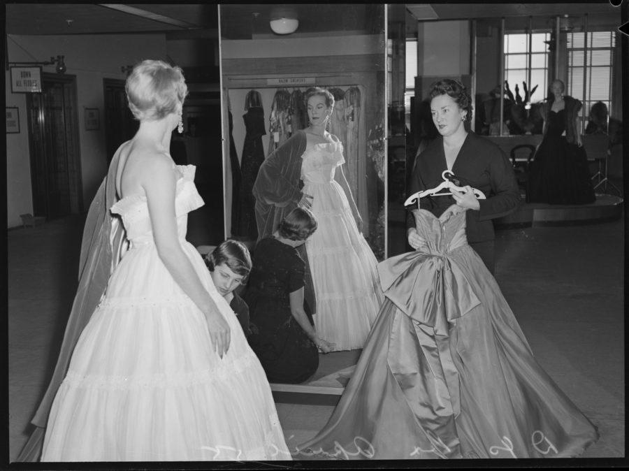 A black and white photograph showing a woman trying a white gown, assisted by two other women.