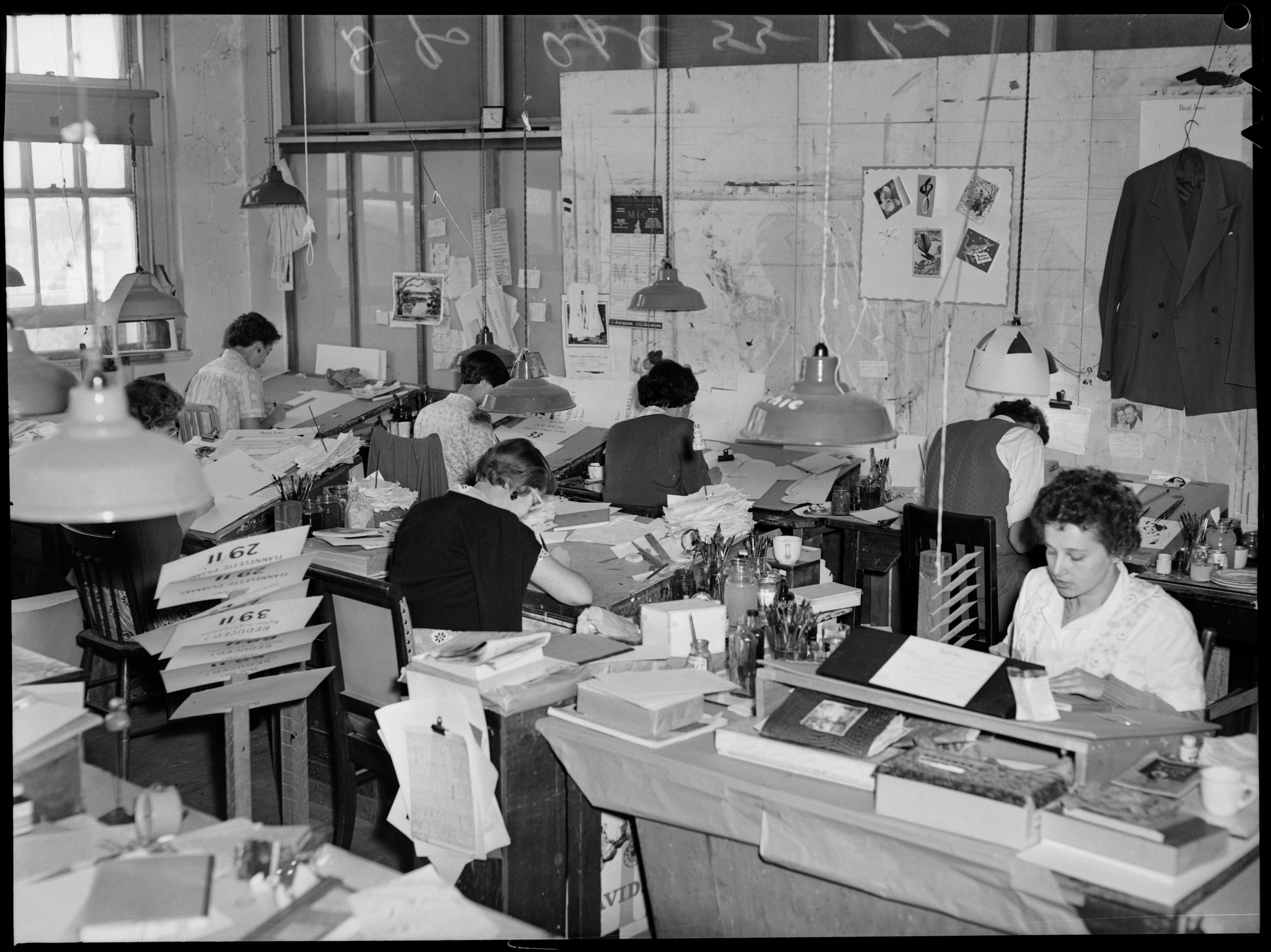 Black and white photo showing several people working at desks. Photographs, plans and papers are pinned to the wall.
