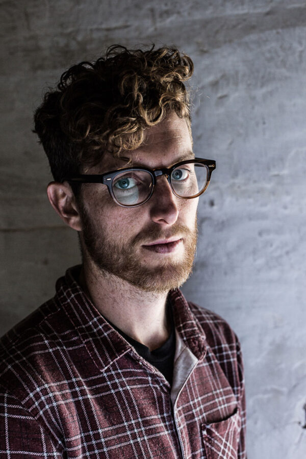 Portrait photograph of a man with curly red hair, a beard and classes wearing a checked shirt.