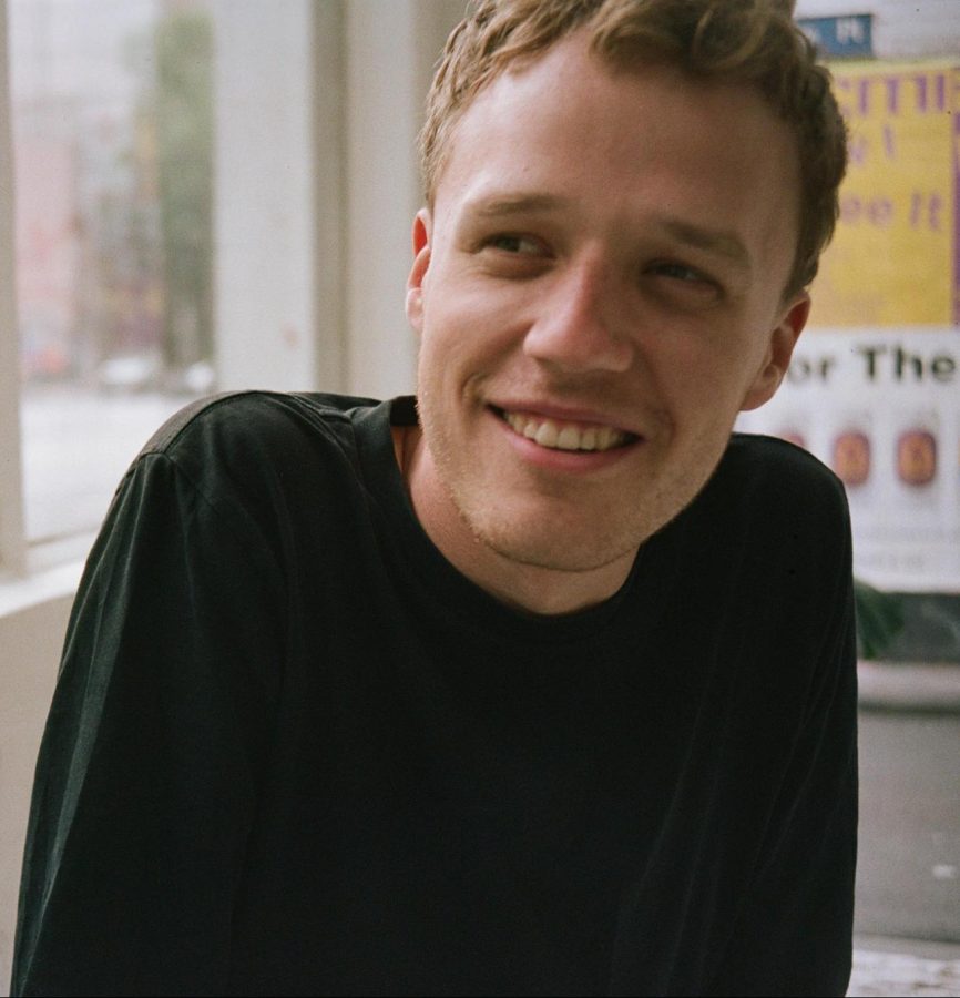 Joshua wears a black jumper and has short cropped blonde hair. He smiles and looks off to the left. His shoulders are slightly hunched as he sits in front of a window.