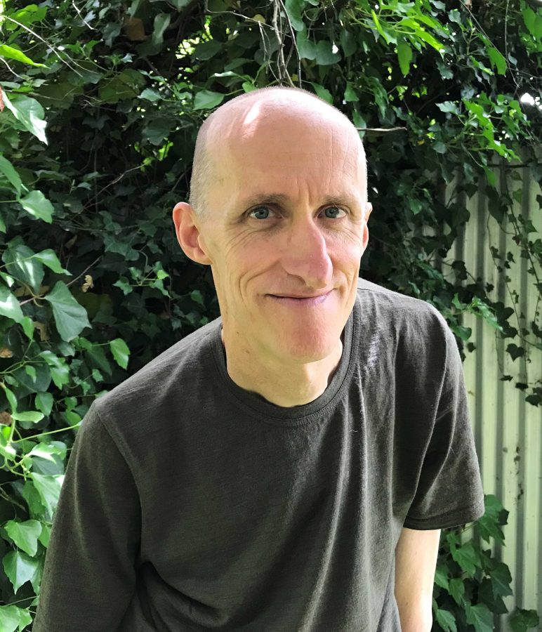 A Caucasian man is wearing a dark green shirt, and sitting in front of a backdrop of lush leaves. He is bald, and smiles slightly as he looks into the camera.