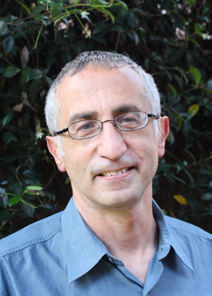 A close-up portrait photo of Abbas El-Zein. He has light grey and black hair, and wears thin black glasses. He is wearing a blue buttoned shirt.