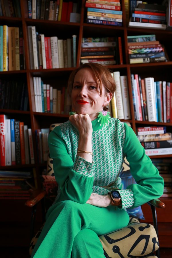 Carody Culver sits cross-legged, and leans her face toward the camera, chin on fist. In front of a large book case. She wears a green dress with long sleeves and a white geometric pattern on the sleeve cuffs and the torso. She has red hair which is tied back, and red lipstick.